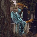 A statue of an angel with light teal patina sits atop a headstone in a cemetery, head downcast.