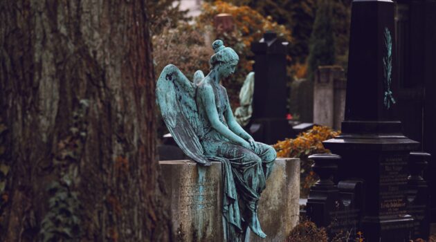A statue of an angel with light teal patina sits atop a headstone in a cemetery, head downcast.