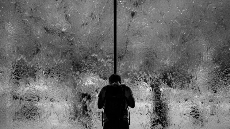 A person stands on one side of a large sheet of ice covered glass, head downcast. Shapes of people are indistinct on the other side.