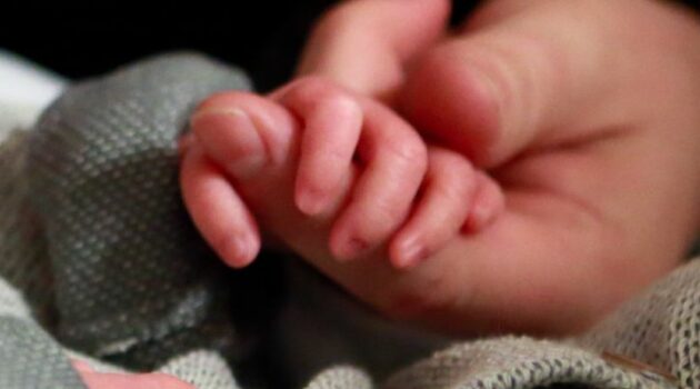 The perfect fingers of a tiny baby boy wrap around an adults left index finger.
