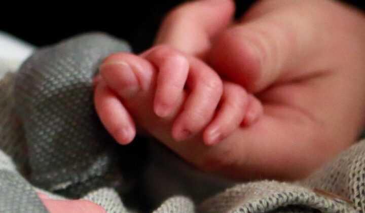 The perfect fingers of a tiny baby boy wrap around an adults left index finger.