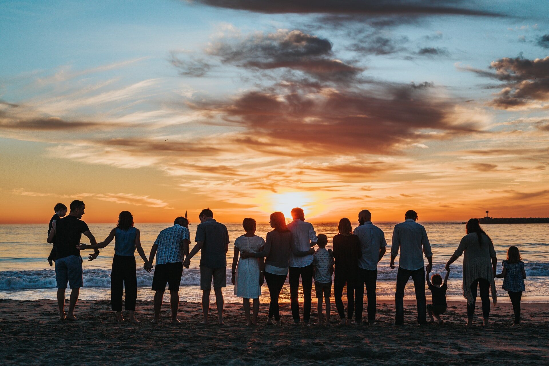 People stand together on a beach, facing the sunset behind the water.